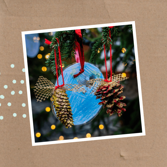 Weihnachtliche Dekoration mit zwei glitzernden Tannenzapfen, die an roten Bändern hängen, vor einem blau bemalten Hintergrund, umgeben von Tannenzweigen und Lichterketten.