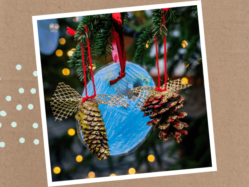 Weihnachtliche Dekoration mit zwei glitzernden Tannenzapfen, die an roten Bändern hängen, vor einem blau bemalten Hintergrund, umgeben von Tannenzweigen und Lichterketten.